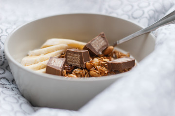 The bowl with granola, banana and chocolate bar. Healthy breakfast in a bed. Close-up of the plate with breakfast