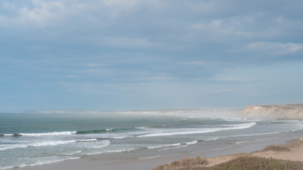 Landscape of the ocean coast in pastel colors. Sandy shore surrounded by cliffs, ocean with waves, sky with clouds.