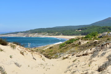 Bay with wild beach and forest, view from sand dunes. Turquoise sea with waves, blue sky. Galicia, Spain.