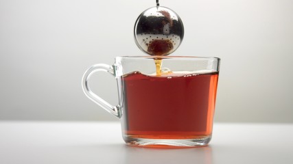 Closeup of transparent cup with brewed tea and a infuser tea spoon lifted out and drops of tea falling into the cup