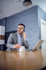 Attractive caucasian businessman sitting at home on dining table, talking on the phone and holding mug with fresh morning coffee. In table is laptop.