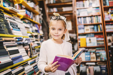 Adorable curious caucasian little girl with eyeglasses standing in bookstore and holding interesting book while looking at camera. All around are books on shelves.