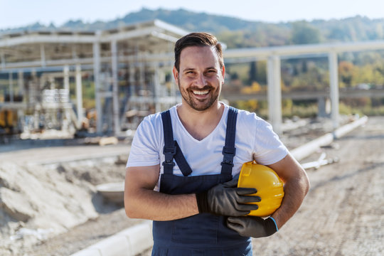 Smiling handsome unshaven caucasian worker in overall standing at refinery with helmet under armpit and looking at camera.