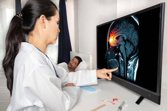 Asian woman doctor touching a screen with x-ray of brain and skull skeleton and showing to a patient man. Headache, trauma and migraine concept. Mask and drugs medicine on the desk.