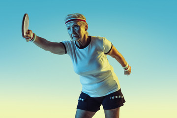Fototapeta premium Senior woman in sportwear training in table tennis on gradient background, neon light. Female model in great shape stays active. Concept of sport, activity, movement, wellbeing, confidence. Copyspace.