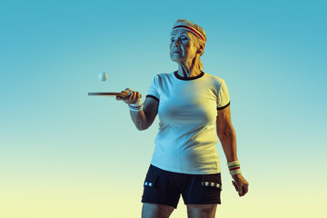 Senior woman in sportwear training in table tennis on gradient background, neon light. Female model in great shape stays active. Concept of sport, activity, movement, wellbeing, confidence. Copyspace.