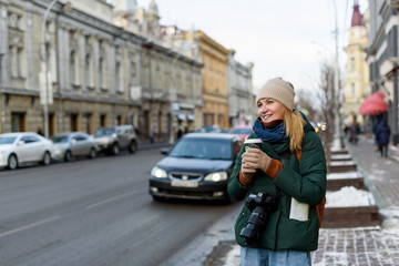 Naklejka na ściany i meble Girl in winter clothes with camera and map in the historical center of the city is holding a cup of coffee on the street