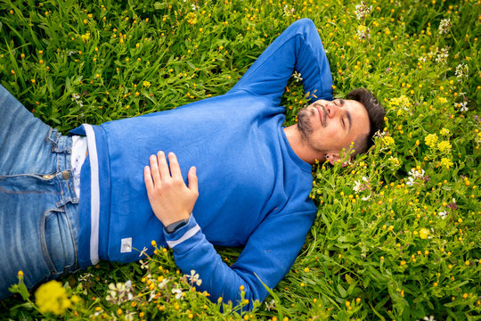 hombre joven y guapo tumbado en un campo de flores, sonr&iacute;e y disfruta de la naturaleza mientras el sol le da en la cara