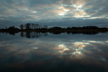 Reflection of clouds in calm water, trees on the horizon, evening view