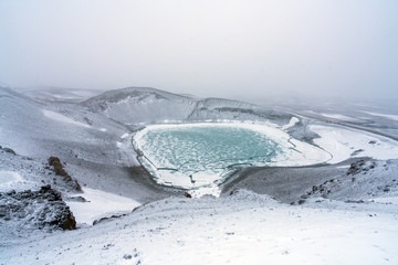 Fototapeta na wymiar Ovelooking ice covered Viti volcanic crater near Krafla geothermal area in Iceland. Foggy and snow covered landscape with frozen green lake at the bottom. Volcano, energy and weather concept.