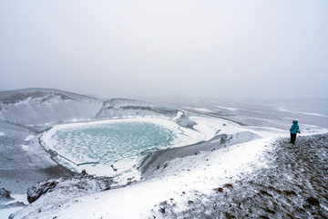 Fototapeta na wymiar Person with blue jacket ovelooking ice covered Viti volcanic crater near Krafla geothermal area in Iceland. Foggy and snow covered landscape with frozen green lake at the bottom. 