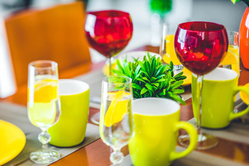 Wooden table. Yellow plates. Mugs. Water in glasses. Wine glasses. Green flower.