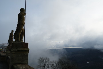 Statues of two Warriors with Speers looking over to the hills surrounding the Hohenzollern castle covered in mist sand clouds in Bisingen Germany. The warriors or guardians are seen in a silhouette. 