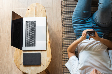 Woman use a smartphone while lying on a sofa with a laptop and ereader on a table. Studying online,...