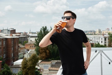 Happy man in black shirt and eyewear drinking beer at bar or pub on roof, copy space