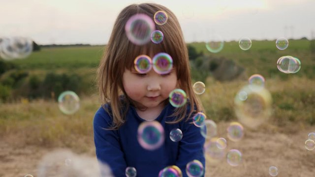 Cute little girl playfully catching soap bubbles outdoors, slow motion shot, soft focus