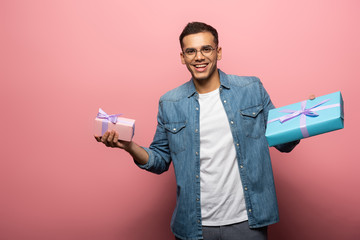 Smiling young man looking at camera while holding gift boxes on pink background