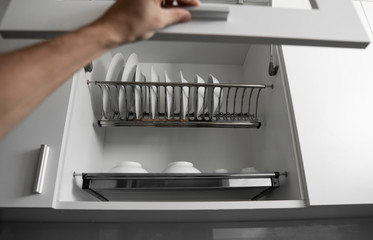 Hand holds the door of the dryer with utensils. Dish drying metal rack with white clean plates. Traditional comfortable kitchen. Open white dish draining closet with wet dishes of plates, bowls.