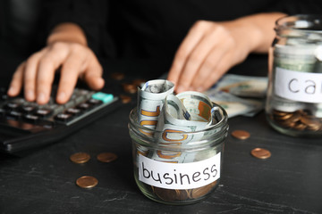 Woman using calculator near glass jar with money and tag BUSINESS on black table, closeup