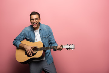 Young laughing man looking at camera while playing on acoustic guitar on pink background