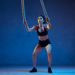 Caucasian young female athlete practicing on blue studio background in neon light. Sportive model training her upper body with ropes. Body building, healthy lifestyle, beauty and action concept.