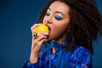 african american woman with makeup eating tasty lemon isolated on blue