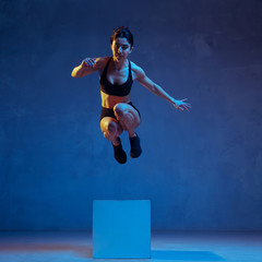 Caucasian young female athlete practicing on blue studio background in neon light. Close up of sportive model jumping on box. Body building, healthy lifestyle, beauty and action concept. Emotional.