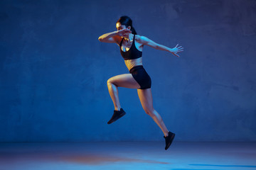 Naklejka na ściany i meble Caucasian young female athlete practicing on blue studio background in neon light. Close up of sportive model jumping high, running. Body building, healthy lifestyle, beauty and action concept.