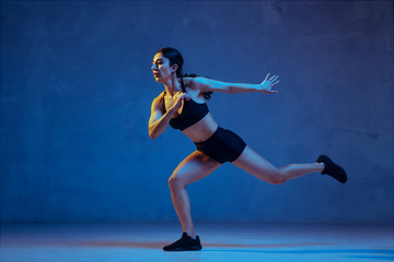 Fototapeta na wymiar Caucasian young female athlete practicing on blue studio background in neon light. Close up of sportive model jumping high, running. Body building, healthy lifestyle, beauty and action concept.