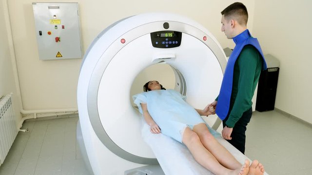 Beautiful female patient lying on back on CT or MRI scan machine in hospital. Handsome man holding her hand comforting. 4K