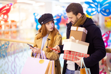 redhaired caucasian woman with shopping bags show something in smartphone to bearded man holding gifts, discuss shopping inside of mall