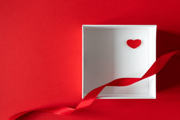 Small wooden heart in white gift box and swirl satin ribbon on red background with copy space, top view. Valentine's day present, flat lay