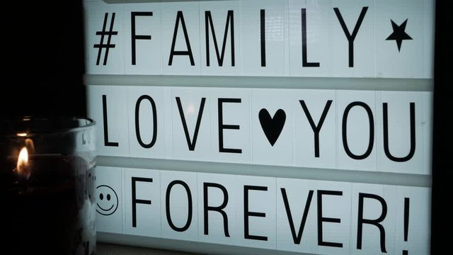 A lit glass candle in front of a billboard with the text Family Love You Forever, a hashtag and some symbols (a heart, a star, a smiley face).
