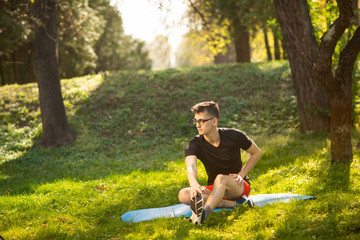 Young man in glasses training yoga outdoors. Sporty guy makes relaxing exercise on a blue yoga mat, in park. Copy space