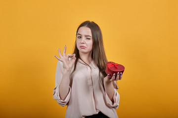 Attractive caucasian young woman keeping red box, doing okay gesture, winks wearing fashion pastel shirt isolated on orange background in studio. People sincere emotions, lifestyle concept.