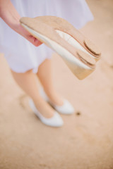 The girl changes her white shoes on the beach and shakes off her feet from the sand