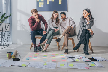 attractive businesswoman pointing with hand at sticky notes on floor near multicultural coworkers