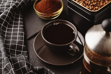 A black coffee Cup,a checkered towel, a metal jar of roasted coffee beans on a dark concrete table