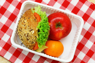 Mixed grain roll with cheese, lettuce and tomato, red apple and mandarine in lunch box on red and white checkered cloth