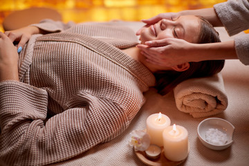 Young girl having massage on her face in spa salon. Wearing bathrobe, candles near her. Beauty treatment
