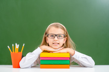 Smiling young girl wearing a eyeglasses sits with books near empty green chalkboard