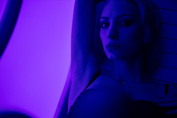 Young blonde woman lies in dark bra with hand up. Photo session on a neon background