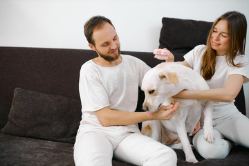 young caucasian couple in white clothes play with friendly white dog, their pet at home, sitting on sofa. people and animals concept