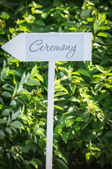 A sign in the form of an arrow on which the ceremony is written pointing towards the wedding event. On a white column on a background of green leaves on a sunny day.