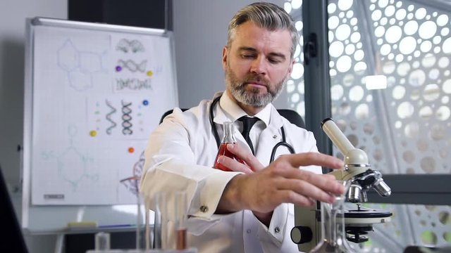 Close view of likable high-skilled gray-haired male doctor which wearing white gown and stethoscope exploring the state of the chemical in the test tube