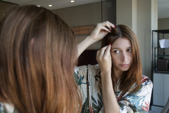 Portrait of a beautiful young woman examining her scalp and hair in front of the mirror, hair roots, color, grey hair, hair loss or dry scalp problem