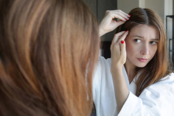 Naklejka premium Portrait of a beautiful young woman examining her scalp and hair in front of the mirror, hair roots, color, grey hair, hair loss or dry scalp problem