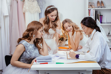 friendly good-looking tailor sit with models on table in office, young girls in wedding dresses made by professional designer, she consultate them and have conversation