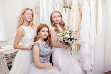 Fototapeta na wymiar three young romantic ladies in wedding dresses, future brides, going to be married, posing together at camera among dresses, holding flowers in hands