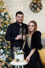 An attractive brunette girl and man with beard banging glasses of champagne at Christmas time. Gender reveal cake with tangerines on baby shower party. Pregnant family, mom and dad in black family loo